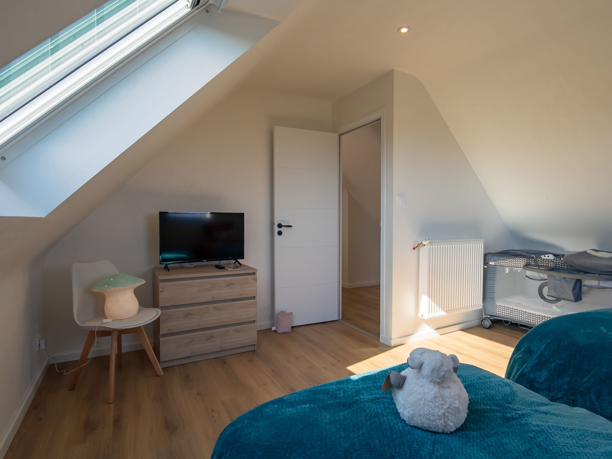 Een moderne, goed verlichte kamer met een schuine kap, een televisie op een houten kast, een deur, een radiator en een bed met een pluche speelgoed.