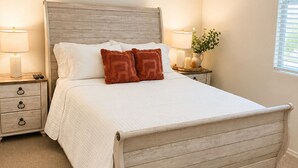 A bedroom with a wooden bed, two bedside tables with lamps, a nightstand, and a window with blinds.