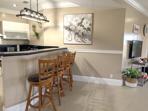 A kitchen with a bar area, wooden chairs, a painting, and a TV on a stand.