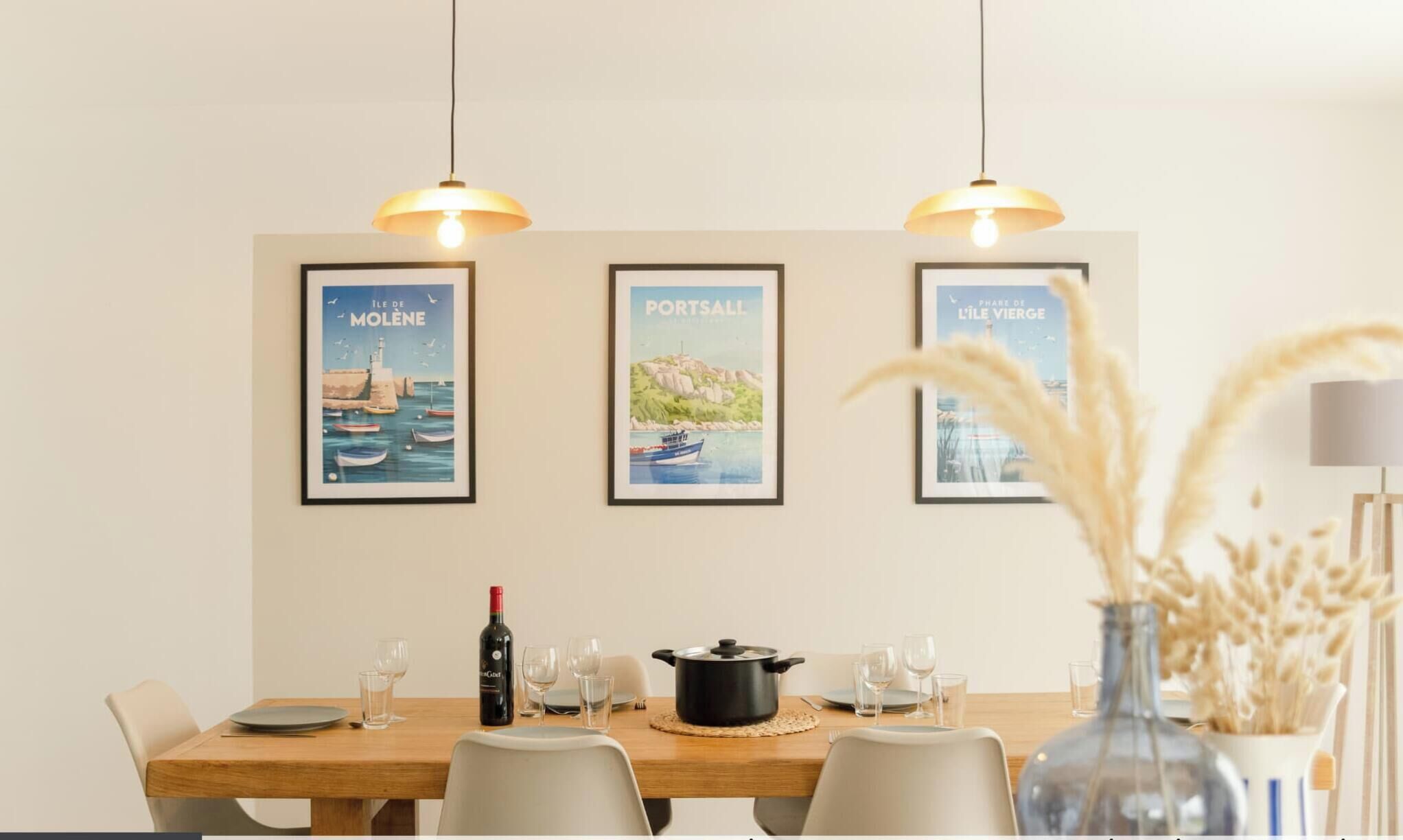 Un comedor con una mesa de madera dispuesta para cuatro, una botella de vino y tres carteles enmarcados en la pared.