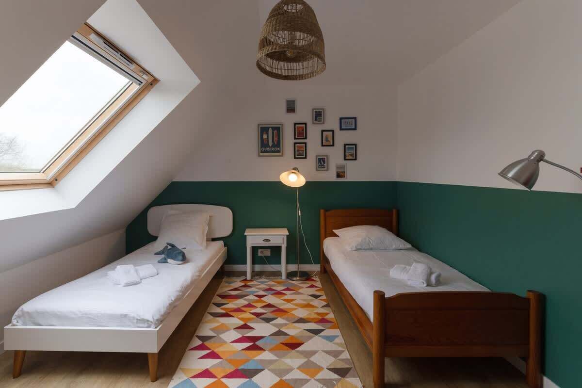 A small, well-lit room with two single beds, a small table, a lamp, and a wall with framed pictures.