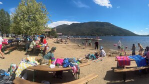 A beach scene with picnic tables, people relaxing, and a lake with boats.