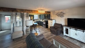 A spacious living room with a sofa, a coffee table, and a TV on a cabinet. There is a kitchen area with a dining table and chairs.