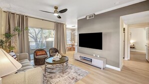 Un salon moderne doté d’un téléviseur à écran plat, de sièges confortables, d’un ventilateur de plafond et offrant une vue dégagée grâce à de grandes fenêtres.