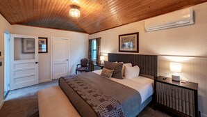 Un dormitorio con techo de madera, una cama con colcha estampada, una mesita de noche con lámpara y un cuadro en la pared.