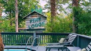 Terraza de madera con sillones de descanso y piscina, rodeada de árboles. Un cartel dice 'Pine Lodge No Vacancy'.