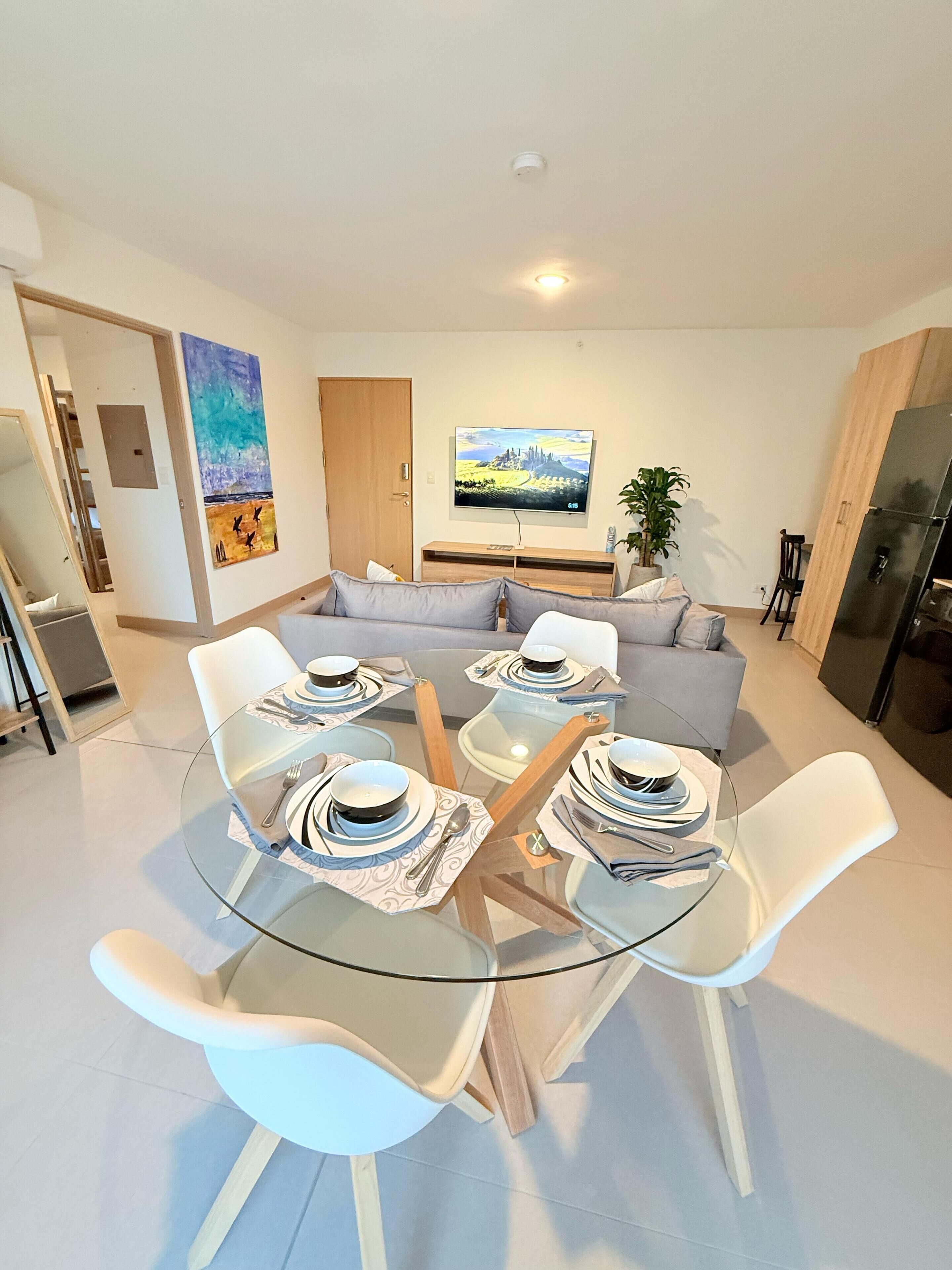 A modern living room with a glass dining table set for four, a grey sofa, a flat-screen TV, and a wooden cabinet.