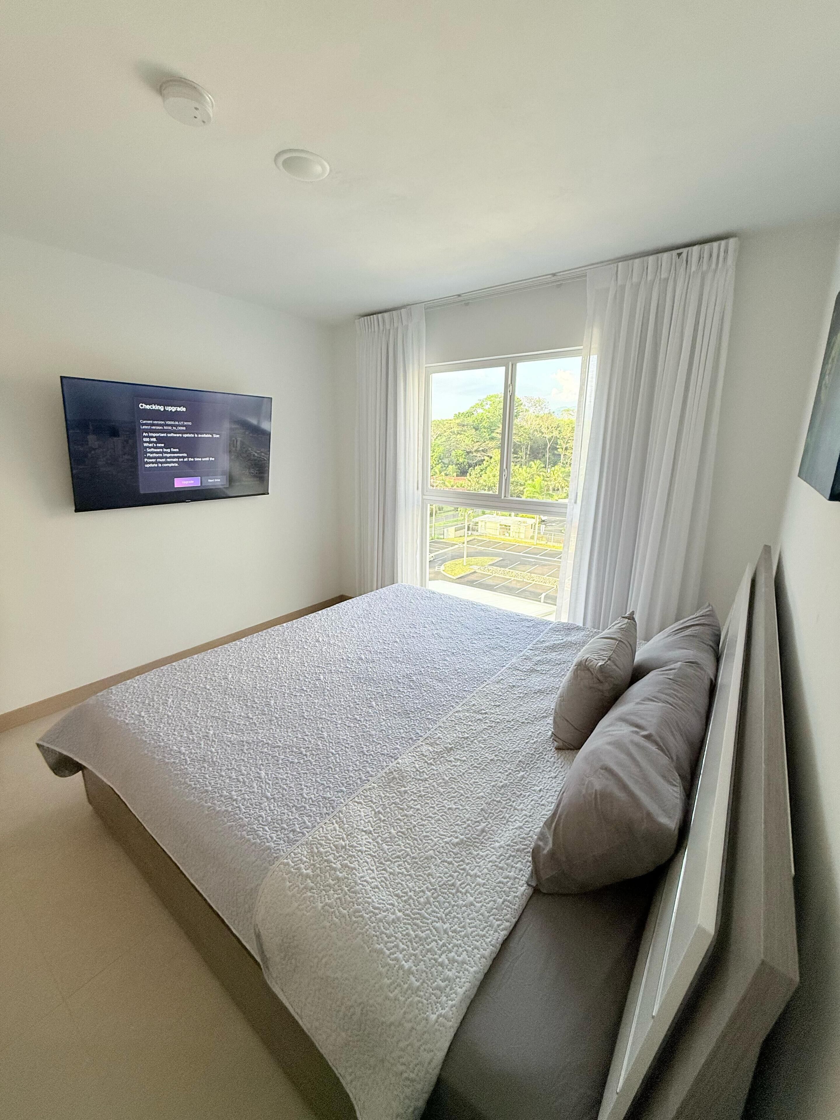 A bedroom with a large bed, a flat-screen TV mounted on the wall, and a window with curtains.