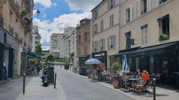 Eine Kopfsteinpflasterstraße gesäumt von Cafés und Restaurants, mit Außenplätzen und einem Roller, der am Straßenrand steht.