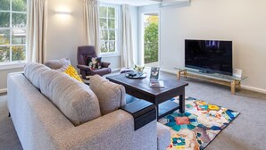 A modern living room with a grey sofa, a dark coffee table, a TV on a glass stand, and a window with curtains. - Fendalton Comfort Stylish and Convenient with Carpark (Christchurch)