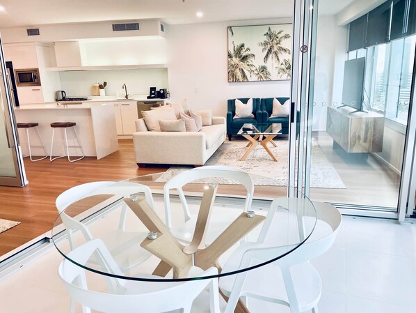 A modern living space with a glass dining table, white chairs, a sofa, and a kitchen area. - Q Tower - Deluxe Ocean View Apartment with King Bed & Spa (Surfers Paradise)