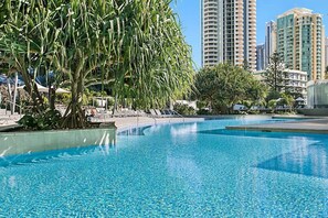 A swimming pool with clear blue water, surrounded by palm trees and lounge chairs. Tall buildings are visible in the background.
