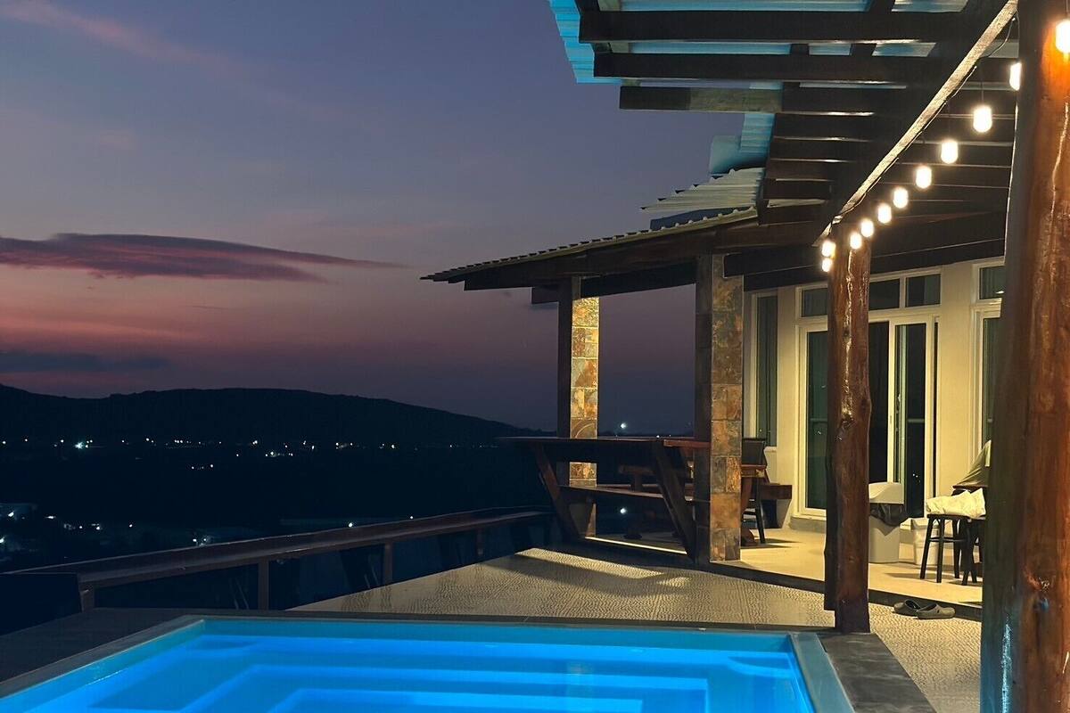 Ein Pool auf dem Dach mit geometrischem Design, eine Essecke mit Außenbestuhlung und Blick auf die Abendstadt.