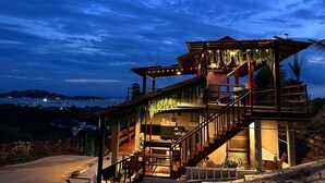 A multi-level wooden structure with outdoor seating and a staircase, overlooking a coastal area at dusk.