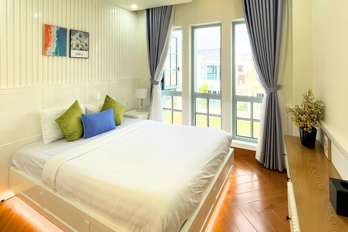 Ein modernes Schlafzimmer mit einem großen Bett, einem Nachttisch, einem Fenster mit Vorhängen und Blick ins Freie.