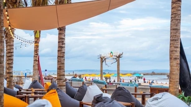 A beachside seating area with wooden tables and chairs, some with cushions. A canopy provides shade, and the beach is visible in the background.