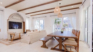 A spacious living room with a large wooden dining table, beige sofas, and a TV mounted on the wall.