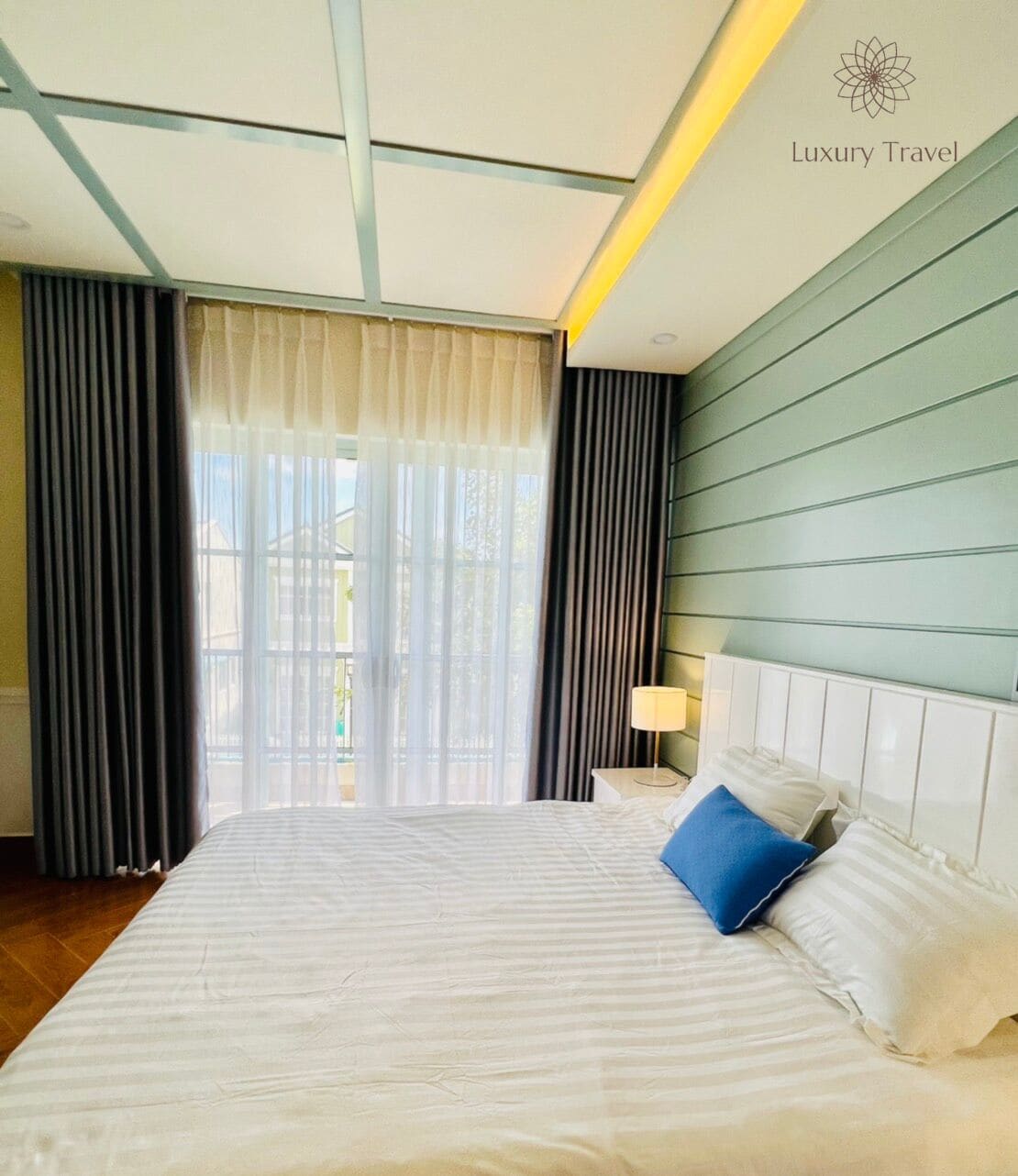 A hotel room with a large bed, a nightstand, a lamp, and a view of the outdoors through the window.