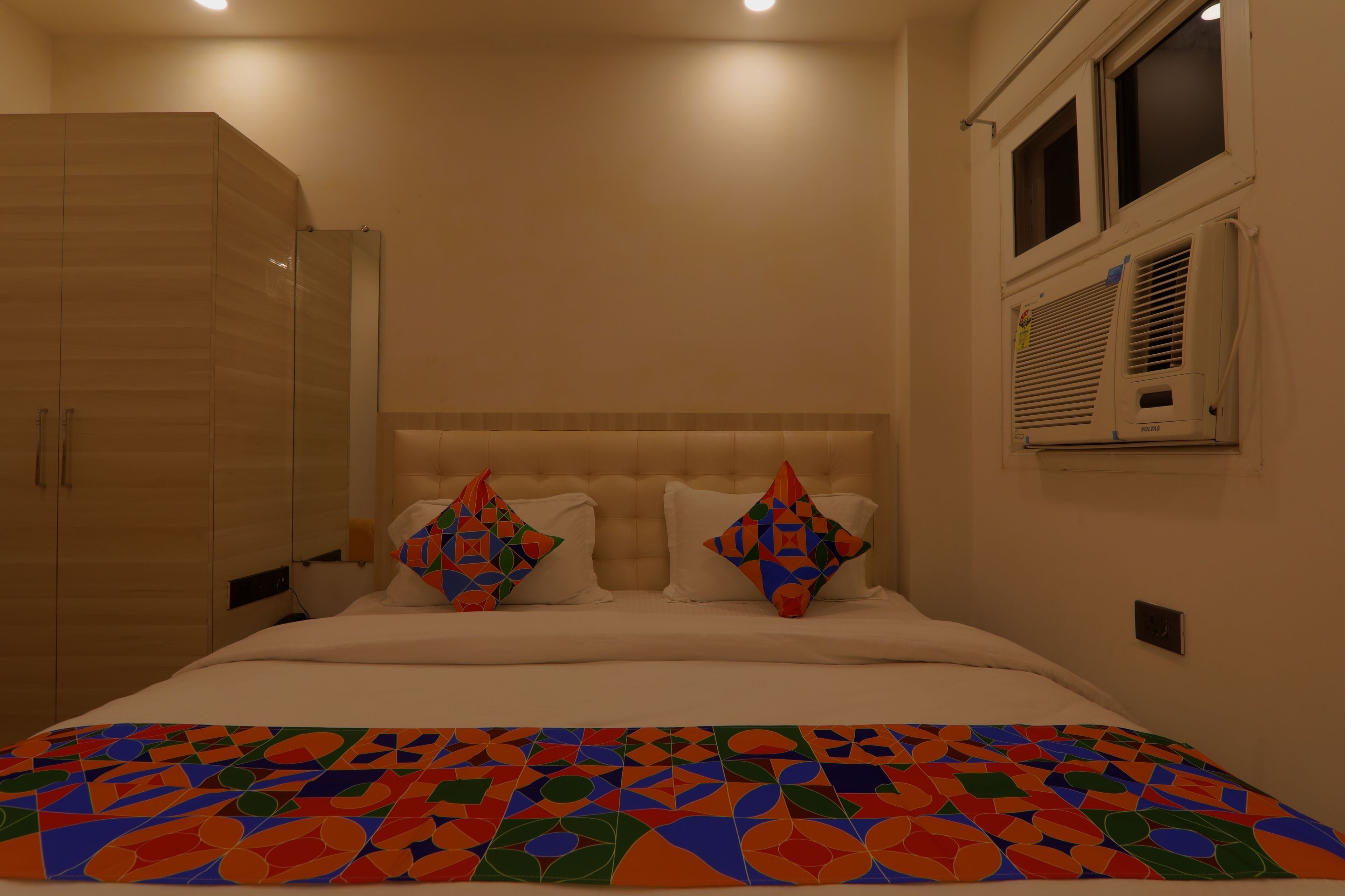 A bedroom with a bed, two pillows, a colorful blanket, a wardrobe, and an air conditioning unit.