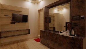 Een moderne badkamer met een grote spiegel, een aan de muur bevestigde televisie en een aanrecht met een waterkoker en kopjes.