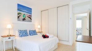 A bedroom with a bed, two bedside tables with lamps, a painting of boats on a beach, and a wardrobe.