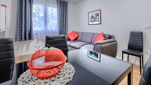 A modern living room with a grey sofa, red cushions, a black chair, a small table with a picture frame, and a window with patterned curtains.