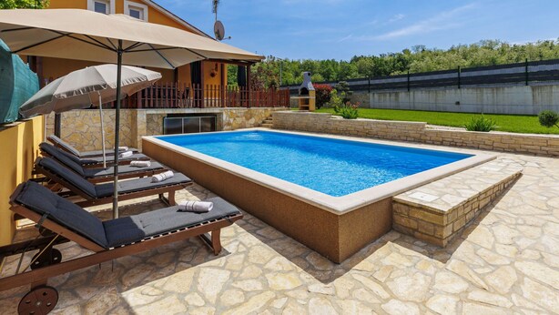 Ein Schwimmbecken mit Liegestühlen und Sonnenschirmen, eine Stegterrasse und eine Steinmauer im Hintergrund.