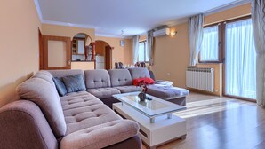 Ein modernes Wohnzimmer mit einem Ecksofa, einem Glastisch und Blick auf die Küche und den Essbereich durch große Fenster.