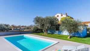 A swimming pool with two white lounge chairs, a blue house, and surrounding greenery.