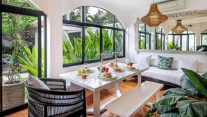 A dining area with a white table and bench, surrounded by large windows offering views of greenery. - Homey 5 BR Villa In Seminyak (Seminyak)