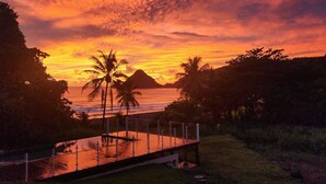 A sunset view with a wooden deck overlooking the ocean, palm trees, and a distant mountain. - F2 M'Bay : Sea & Pool access (Anse Charpentier)