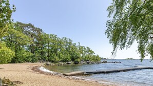 Ein sandiger Strand mit einer Mole, die in ruhiges Wasser führt, umgeben von dichtem Grün.