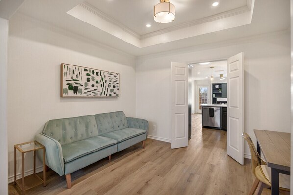 Una sala de estar moderna con un sofá verde claro, una mesita de madera y una cocina visible a través de una puerta abierta.