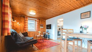 Un acogedor interior de cabaña con techo de madera, una zona de comedor con mesa y sillas, una cocina con estufa y una sala de estar con sofá y alfombra roja.
