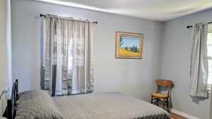 Un dormitorio con cama, una silla, un cuadro en la pared y cortinas.