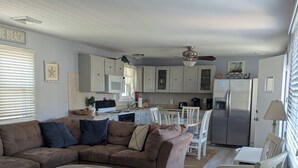 A spacious living room with a sectional sofa, a coffee table, and a patterned rug. A kitchen with white cabinets and a refrigerator is visible in the background. - Charming 3-Bedroom Wildwood Crest Spot: Beach, Bay, and Boardwalk- all so close! (Wildwood)