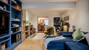 A living room with a fireplace, a blue sofa, a green armchair, and a bookshelf. - Beautifully presented holiday cottage - Stonecrop Cottage (Moreton-in-Marsh)