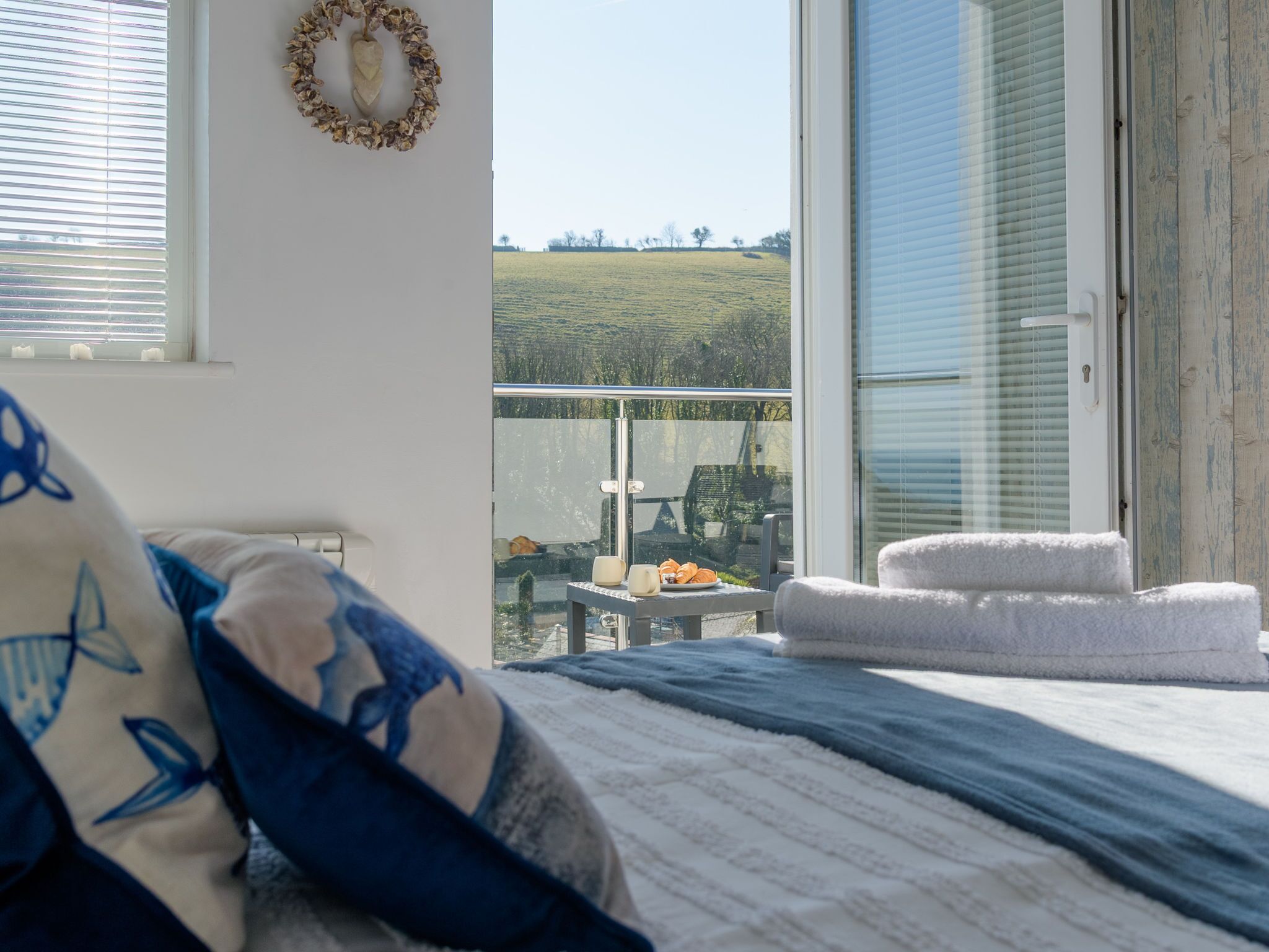 A bedroom with a large bed, a balcony with a table and chairs, and a view of a green field.