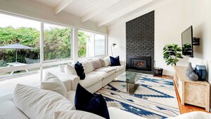 Ein modernes Wohnzimmer mit einem weißen Sofa, einem Glastisch, einem gemusterten Teppich, einem Kamin und einem großen Fenster mit Blick auf Grünflächen.