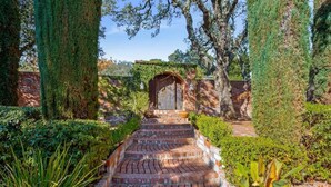 A brick pathway leading to a wooden door with a brick archway, surrounded by lush greenery and tall trees. - The Curated Haven, by Vinifera Homes (Sonoma)