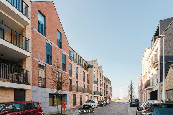 Un moderno edificio de apartamentos con varios balcones, fachada de ladrillo y una fila de coches estacionados en la calle.