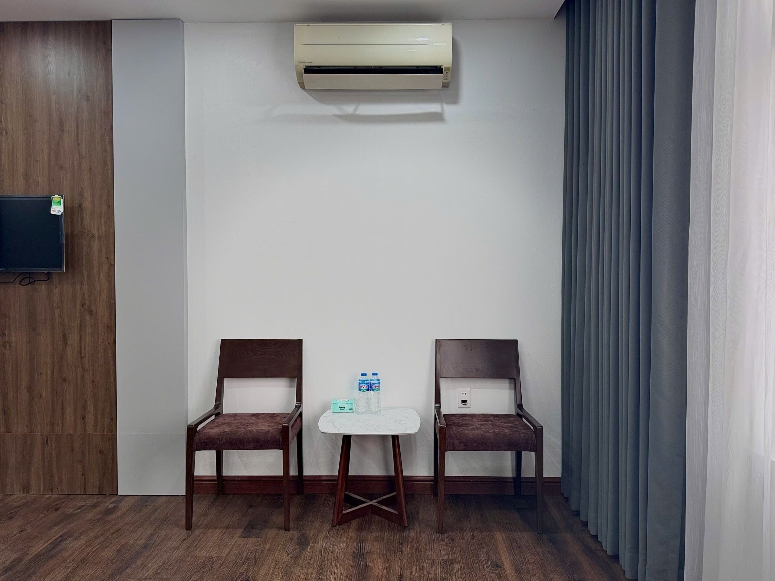 A hotel room with two wooden chairs, a small white table, a flat-screen TV mounted on a wooden panel, and an air conditioning unit on the wall.