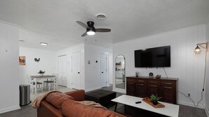A modern living room with a brown leather sofa, a dining area with a white table and chairs, a kitchen with a large mirror, and a mounted TV. - 69 Unit- B (Cartersville)