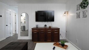 A modern living room with a flat-screen TV mounted on the wall, a wooden cabinet with drawers, a large mirror, and a white table with a remote control and a tray. - 69 Unit- B (Cartersville)