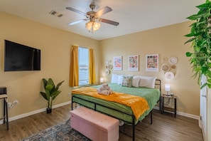 Une chambre à coucher comprenant un lit, un ventilateur de plafond, une télévision, une fenêtre avec des rideaux et des œuvres d’art mural.