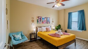 Une chambre à coucher comprenant un lit, une chaise, un ventilateur de plafond, des œuvres d’art accrochées au mur et une fenêtre avec des rideaux.