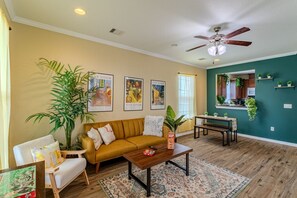 Un salon comprenant un canapé, une table basse et un coin repas avec une table et des chaises. On y trouve également des plantes, un ventilateur de plafond et des œuvres d’art encadrées accrochées aux murs.
