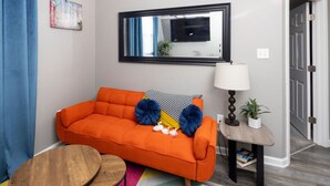 Ein Zimmer mit einem hellen orangefarbenen Sofa, einem kleinen Holztisch, einem farbenfrohen Teppich, einer Lampe und einem Spiegel an der Wand.