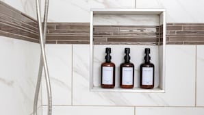 A shower with a handheld showerhead and a flexible hose, mounted on a wall with white marble tiles and a brown decorative border. Below, there is a shelf with three bottles labeled shampoo, conditioner, and body wash. - Modern City Oasis: Pet Friendly | Downtown | Park (Little Rock)