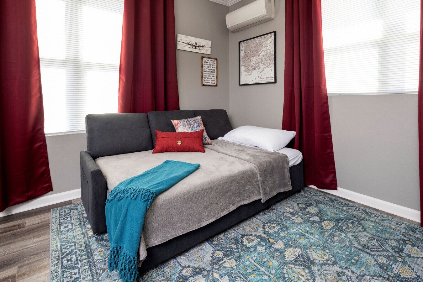 A bedroom with a grey sofa bed, red and white pillows, a blue throw blanket, and a patterned rug. There are two framed pictures on the wall and a window with red curtains.